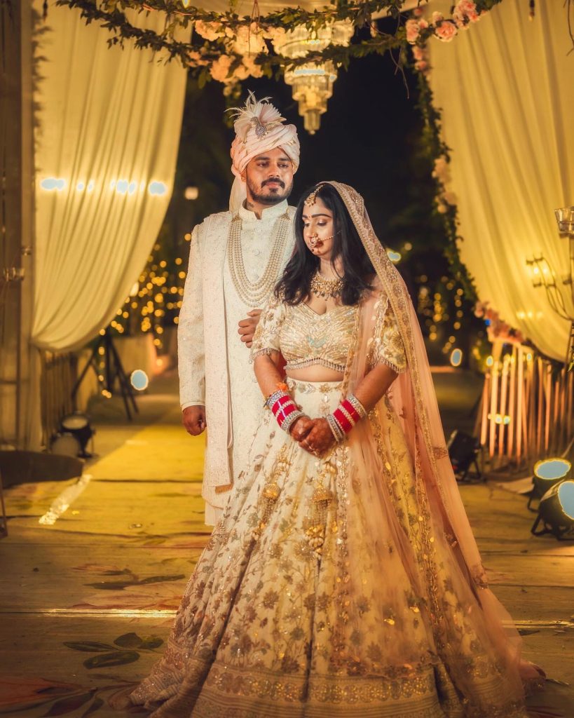 Manika and Kaustubh in their wedding attire beneath ivory drapes and chandeliers in Jim Corbett